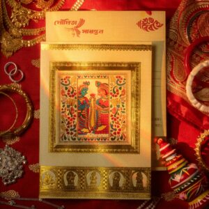 A beautiful traditional Indian wedding card with intricate design surrounded by jewelry on a red fabric.