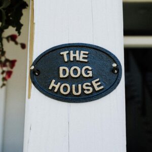 Close-up of a rustic 'The Dog House' sign on a white wooden post with foliage.