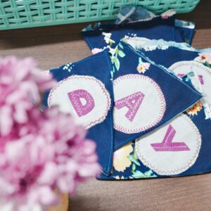 Close-up of vibrant fabric banners with stitched letters on a wooden table, beside pink flowers.