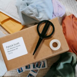 Top view of tape with scissors on carton box with exact address and name among clothes
